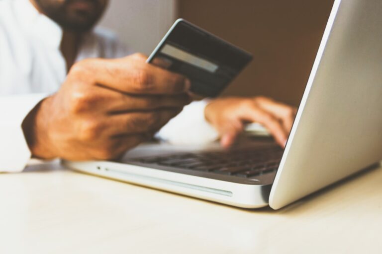 A small business owner at a laptop reviews website payment settings with a smartphone and notepad nearby.